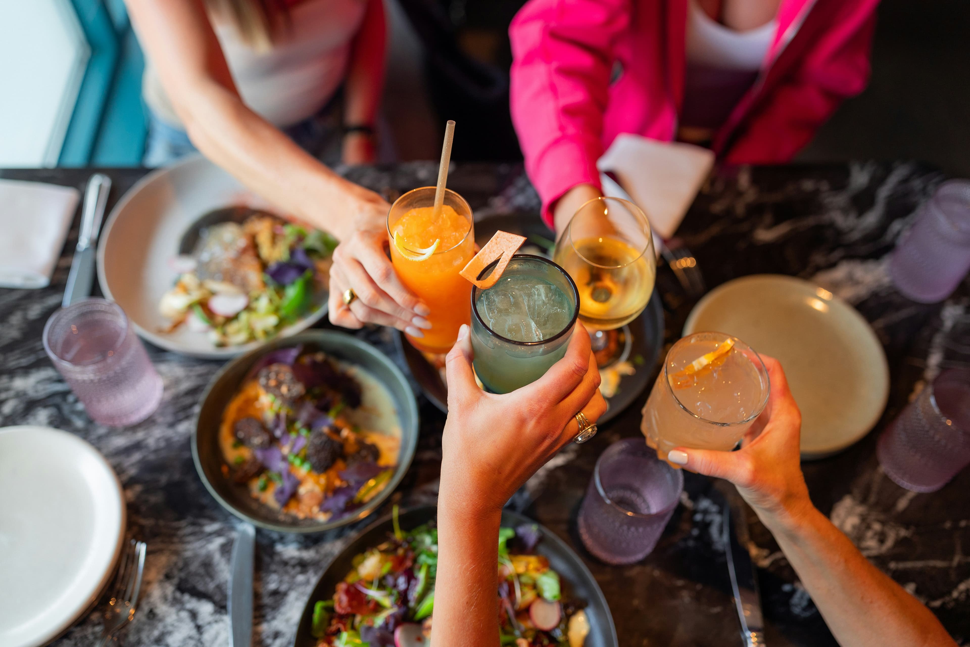 Friends clinking cocktail glasses together over a table of shared plates during happy hour